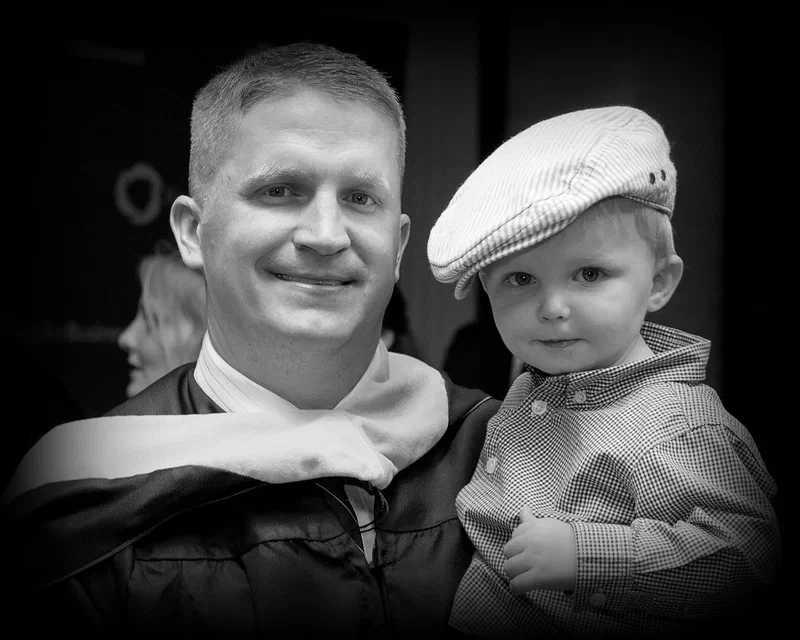 Black-and-white photo of a smiling graduate in academic regalia holding his toddler son, who wears a flat cap.