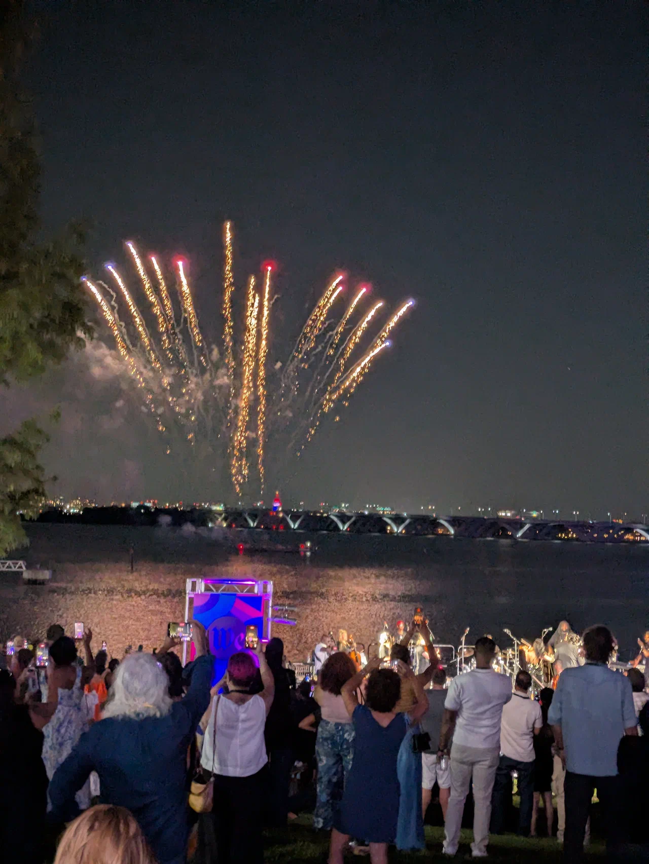 Fireworks over a river at sunset as a crowd stands facing the fireworks and watching.