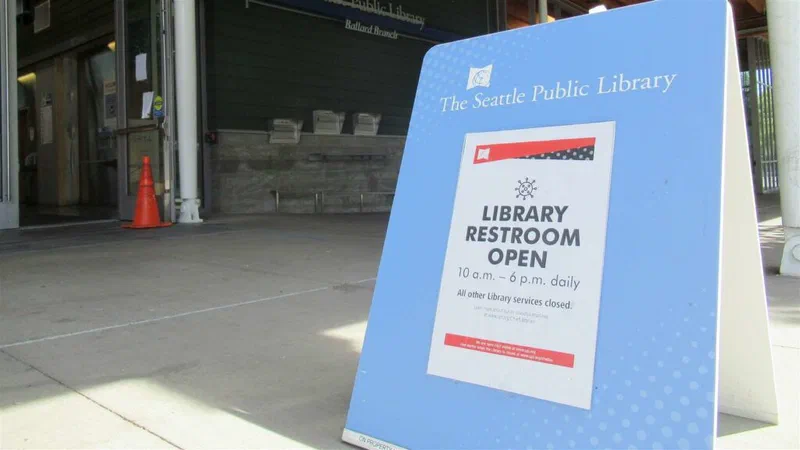 A sign outside the Ballard Branch of The Seattle Public Library indicates that the restroom is open from 10 a.m. to 6 p.m. daily, with all other library services temporarily closed. The sign emphasizes that the funds are not yet spent and encourages community input on how ARPA aid should be used.